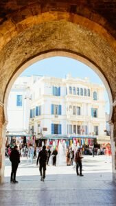 A bustling town square with an archway overlooking a fountain and vibrant stalls.