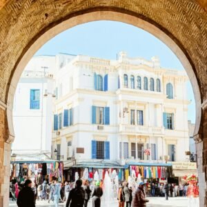 A bustling town square with an archway overlooking a fountain and vibrant stalls.